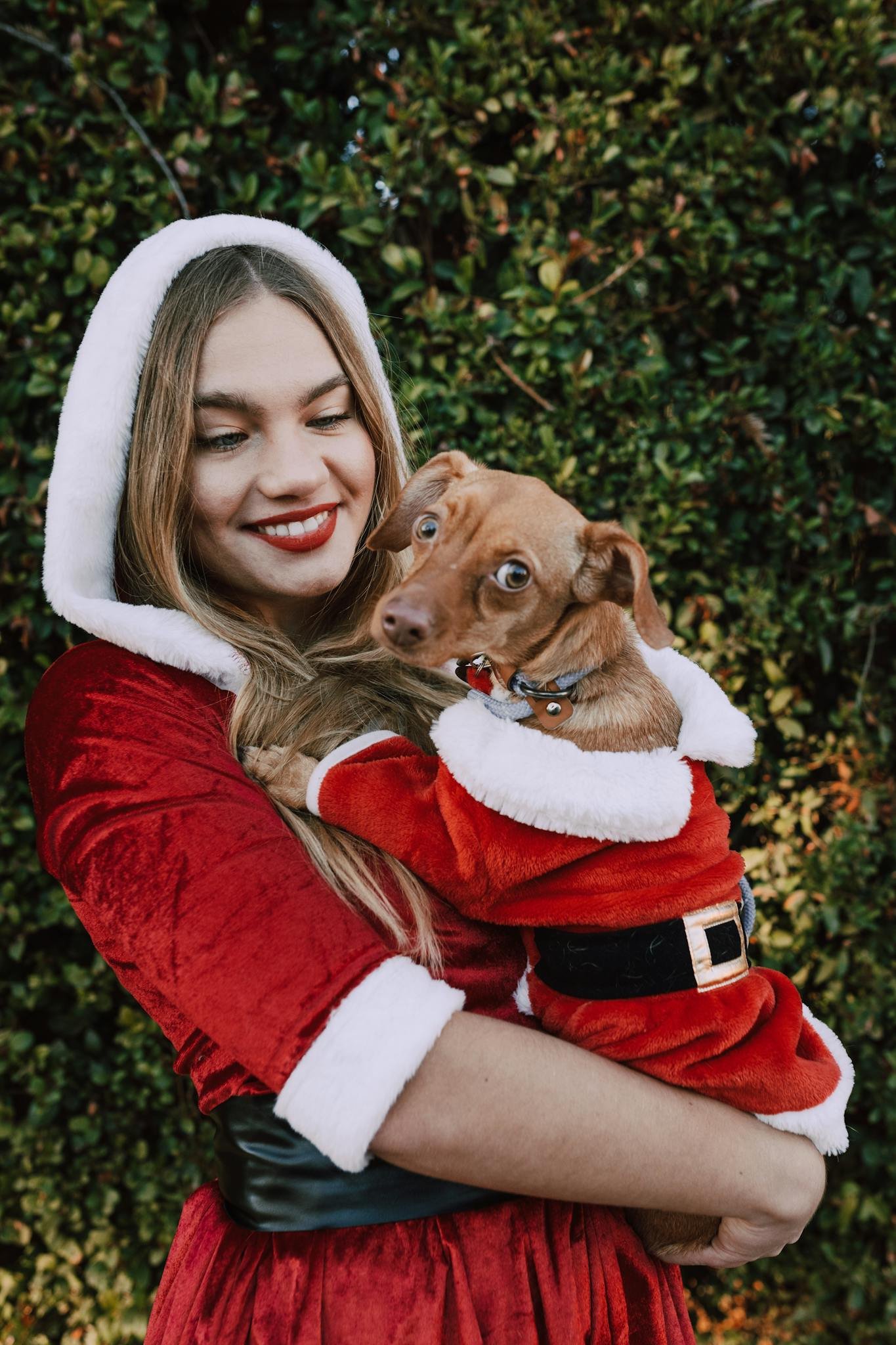 A woman dressed in a Santa costume joyfully holds her dog, also dressed as Santa, in an outdoor setting.